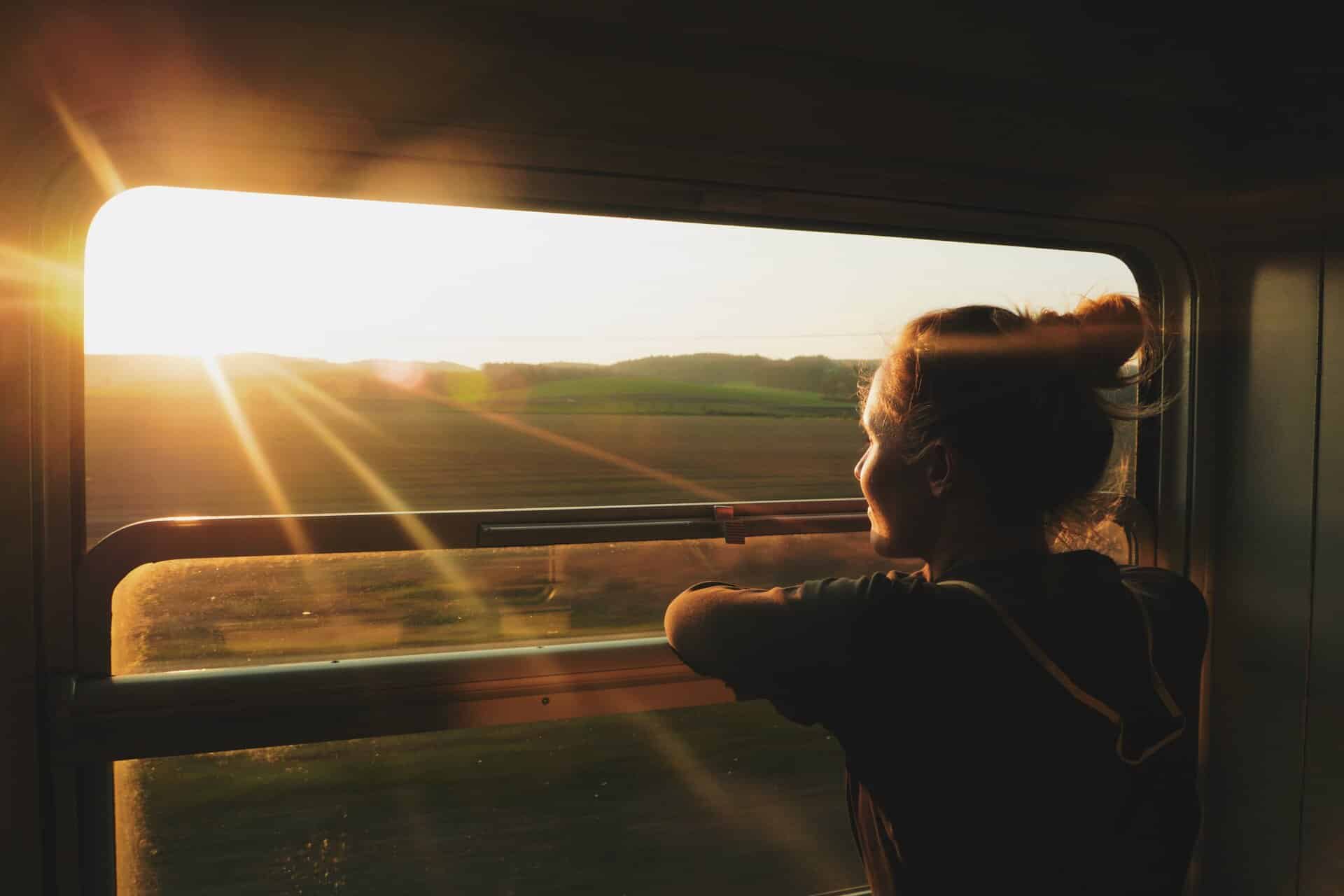 Junge Frau blickt aus dem Zugfenster in eine sonnige Landschaft